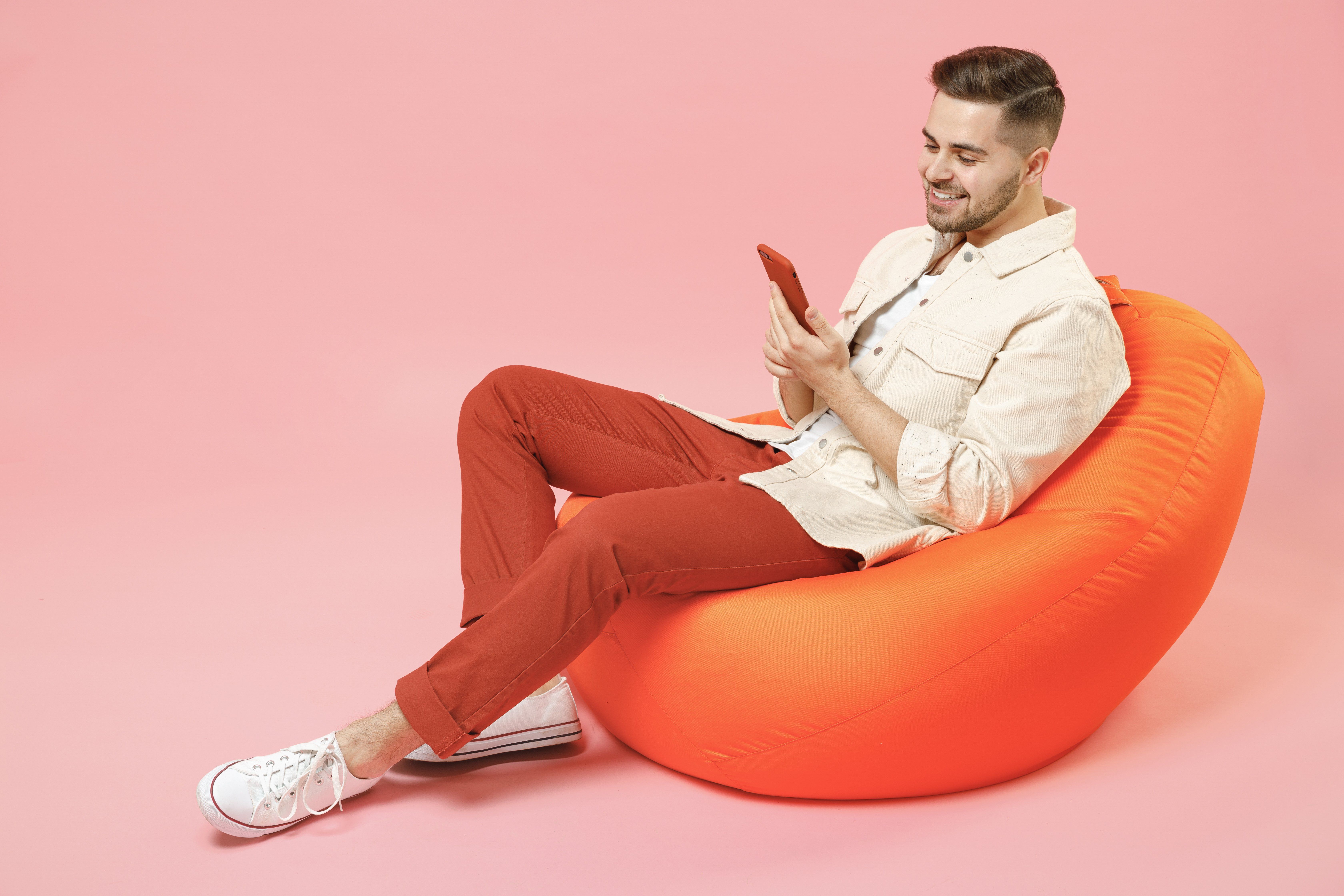Full length young smiling fun man 20s wearing jacket white t-shirt sitting in bean bag chair hold mobile cell phone chatting online in social network isolated on pastel pink color background studio.