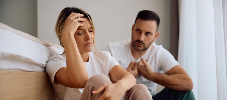 Mid-30s woman sitting by bed, holding her head in frustration as her male partner tries to explain himself.