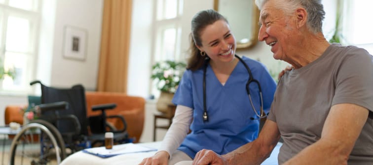 A senior at a nursing home sitting on the bed with a nurse.
