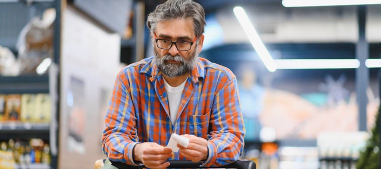Bespectacled man with gray hair and beard leaning on his shopping cart and holding a receipt with a distant look in his eyes.