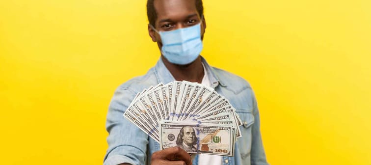 Portrait of joyous wealthy man with surgical medical mask holding out dollar bills at camera, boasting money won in lottery. indoor studio shot isolated on yellow background