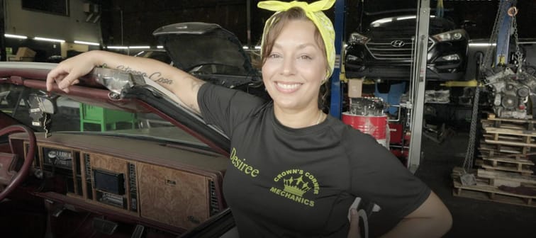 Georgia mom proudly standing and smiling in her auto mechanic shop, in a black company t-shirt and a lemon yellow bandanna tied in a bow on top of her head.