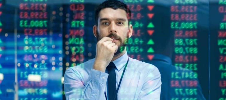 Stock Market Top Trader Looks at Projected Ticker Numbers and Graphs Running, Analysing Data to Make Best Sell. Behind Him Room Full of Screens and Statistics.