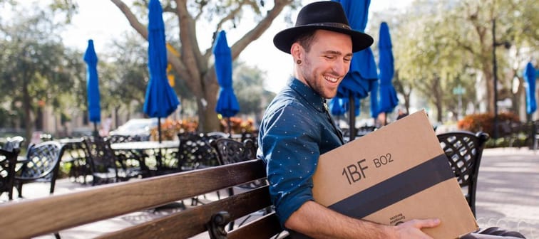 Hipster wearing a wide-brimmed hat sitting on a park bench holding a box from Amazon