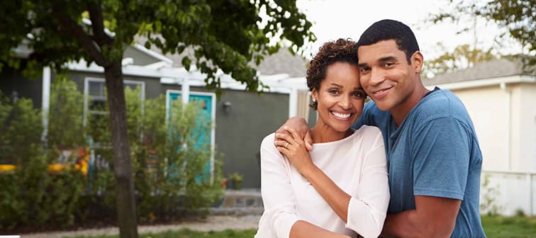 young couple with their arms around each other, smiling in front of home