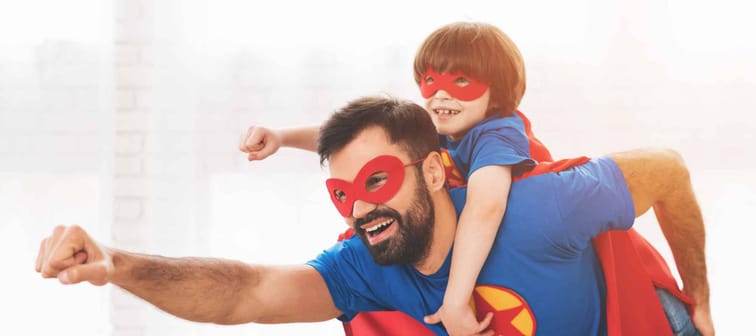 Father and son in the red and blue suits of superheroes. On their faces are masks and they are in raincoats. They are posing in a bright room.
