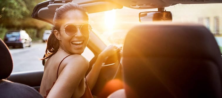 Smiling young woman with blonde hair and sunglasses has handle on the wheel of a car and looks behind her