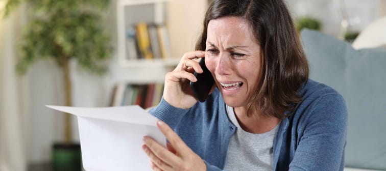 Sad adult woman crying calling on smart phone with letter sitting on the floor at home