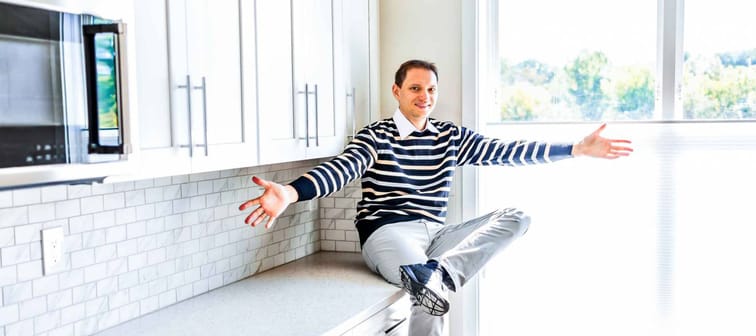 Young man sitting on kitchen countertop with outstretched open arms in clean, modern, white home design