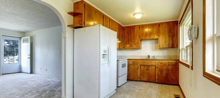 Empty home, older cabinets in the kitchen, beige carpeting in the living room adjacent