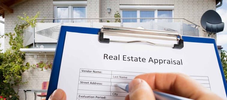 Close up of person's hand holding clipboard with sheet that says "Real estate appraisal" and home in the background