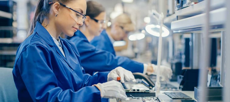 Female Electronics Factory Worker in Blue Work Coat and Protective Glasses is Assembling Laptop's Motherboard with a Screwdriver. High Tech Factory Facility with Multiple Employees.