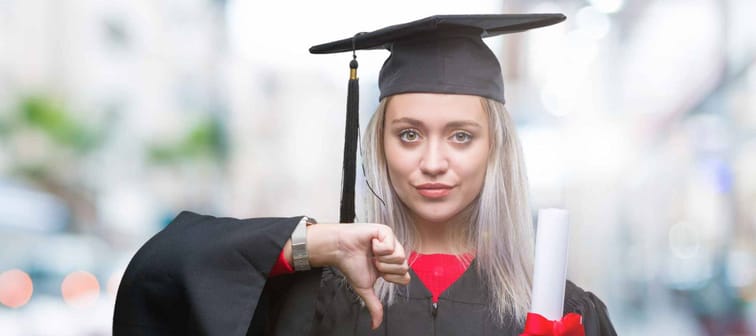 Young blonde woman wearing graduate uniform holding degree over isolated background with angry face, negative sign showing dislike with thumbs down, rejection concept