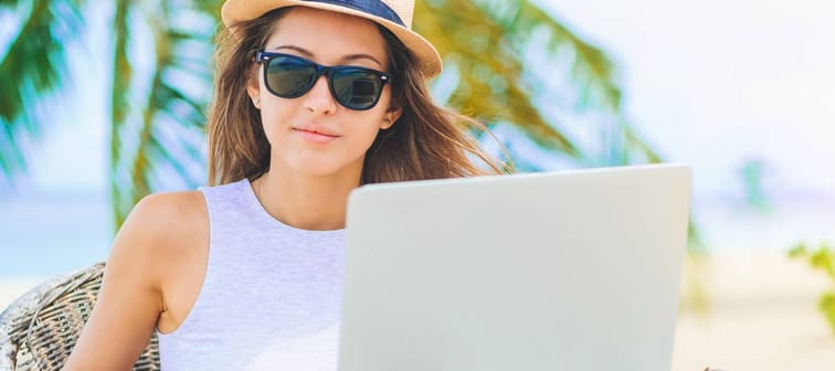 Young attractive woman in sunglasses and dress with hat working in laptop on the beach. Girl Freelancer working