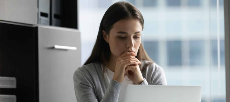 Thoughtful focused businesswoman looking at laptop screen, sitting at desk in modern office, young woman employee worker working on difficult project, pondering strategy, reading email
