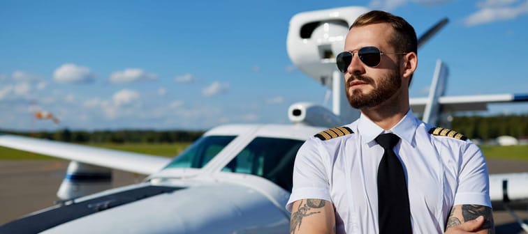 Cool young pilot in sunglasses with tattoos on his hands posing at private air plane on runway. Good summer weather, handsome male model