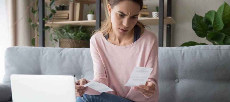 Stressed worried young woman holds receipts, sitting on couch in front of laptop