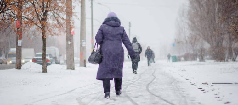 elderly woman in dark coat walks along road that is not snowed off in city. Poor utilities work. Not working snow equipment. Heavy blizzard and snowfall in city.