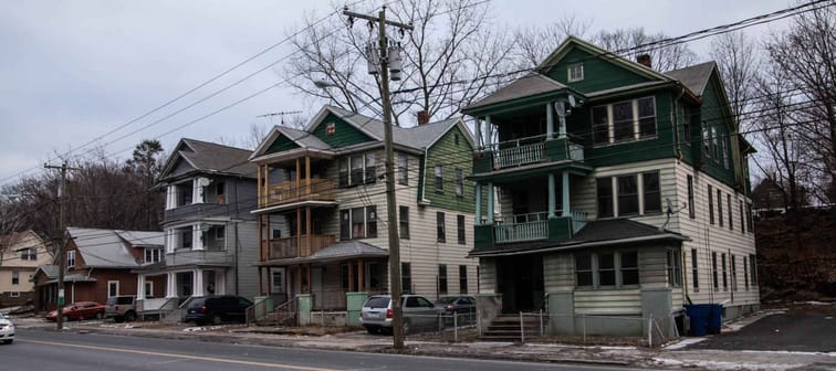 The run-down neighborhood with decrepit buildings surrounded by chain-link fences and old cars parked next to it on a gloomy, grey winter day.