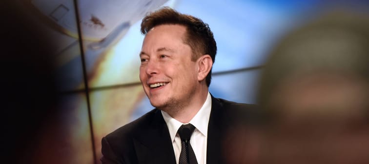 Elon Musk smiles to the side of the camera, wearing a suit