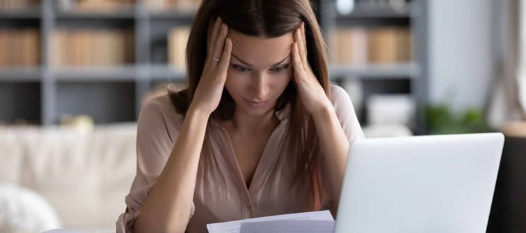 Stressed young woman holding head in hands, looking down at laptop and papers