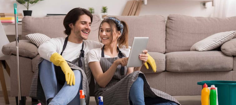 Cheerful Young Couple Relaxing With Digital Tablet After Cleaning Apartment, Ordering Food Delivery Online Or Browsing Internet
