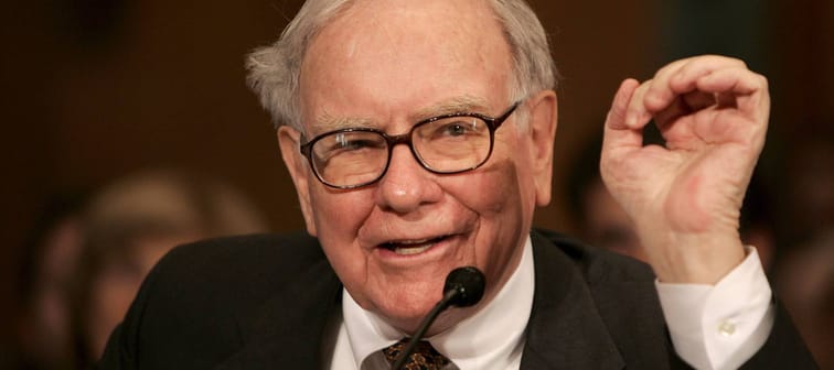 Warren Buffett sits in front of microphone, talking and holding up hand in gesture