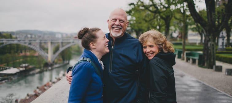 Older couple pose with younger woman on a riverside walk, everyone is smiling and happy