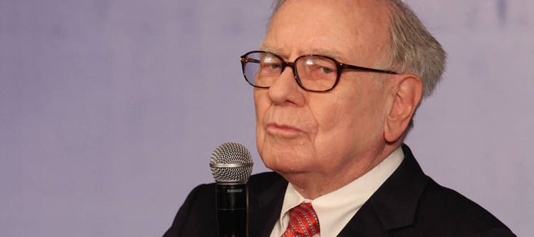 Warren Buffett holds a microphone and looks to the side of the camera with his eyes narrowed