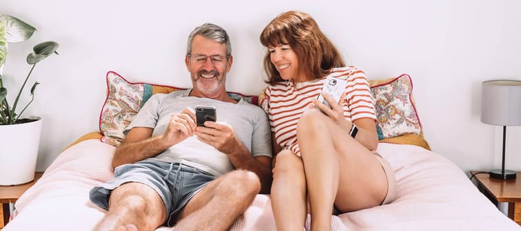 Middle-aged couple using their mobile phones laying on a bed and laughing.