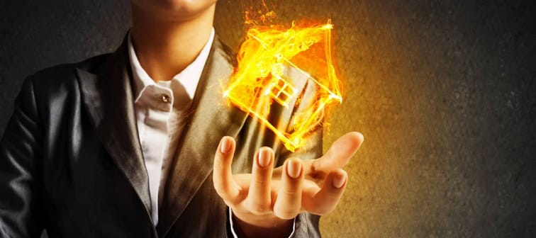 Closeup of businessman in black suit keeping flaming house symbol in hands with dark wall on background.