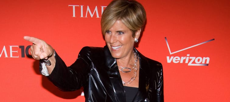 Suze Orman stands in front of a red backdrop with her finger pointing