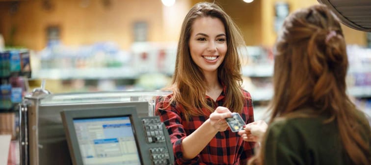 Image of smiling young lady standing in supermarket shop near cashier's desk holding credit card. Looking aside.