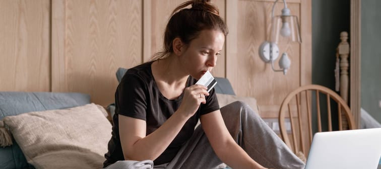 Woman sitting on bed, surrounded by papers and holding a credit card to her lips.