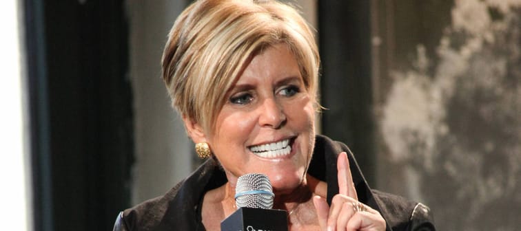 Suze Orman sits on a stage with her mouth open, holding a microphone and talking.