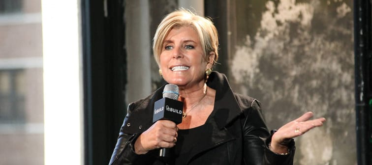 Suze Orman speaks on a stage with a microphone in her hand and a hand making a waving gesture.