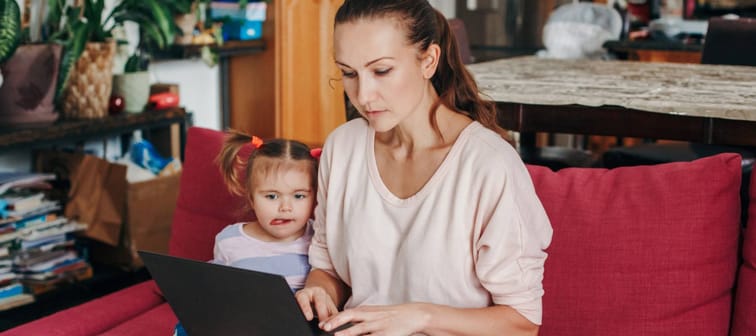 Mother working on laptop with baby girl toddler sitting beside her.