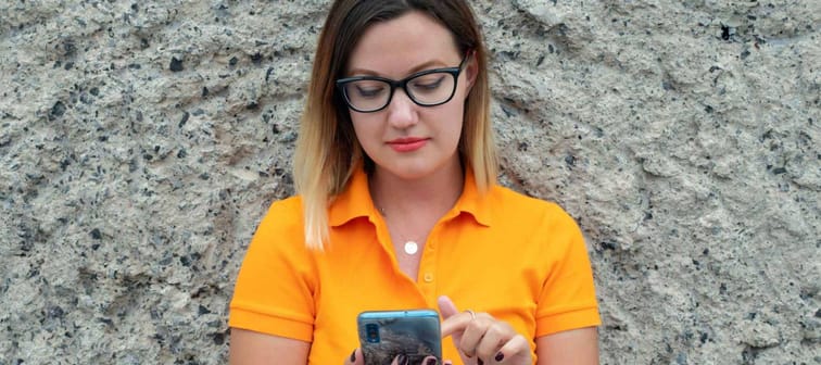 Young millennial woman wearing glasses and yellow polo shirt while checking out her smart phone, looking to see if she received a tax refund.