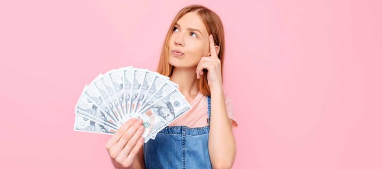 Young thinking attractive girl holding cash dollar bills, looking away, on a pink background