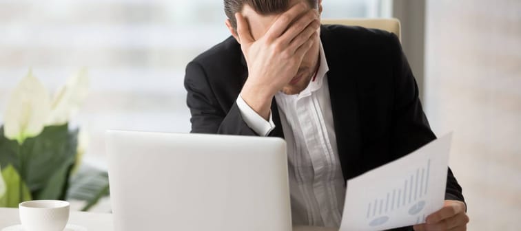 Stressed unhappy young businessman in office in front of laptop, holding financial document and facepalming
