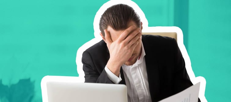 Stressed unhappy young businessman in office in front of laptop, holding financial document and facepalming