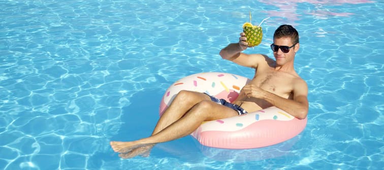 CLOSE UP: Smiling guy sitting on doughnut floatie drinking pineapple cocktail