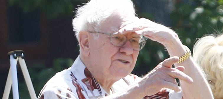 Warren Buffett stands outside, shading eyes from sunlight, pointing finger.