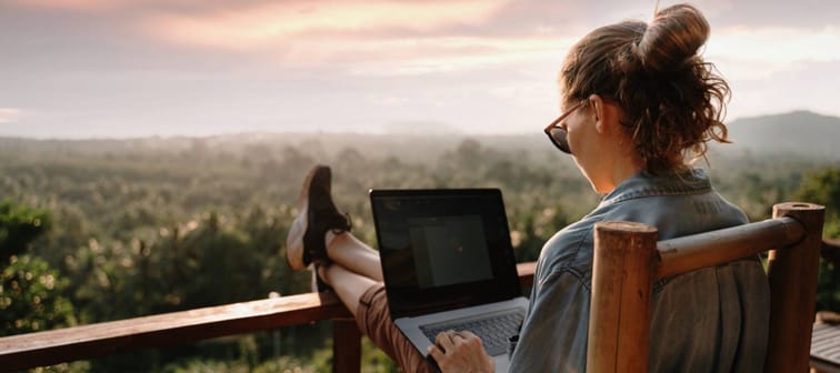 A woman works remotely on her laptop on a porch overlooking a sunset on a beautiful landscape of trees