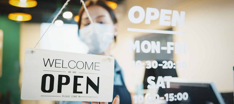 Asian female restaurant owner wear face mask, flip the shop's sign placard from Closed to Open. Business reopen after covid-19 coronavirus pandemic.
