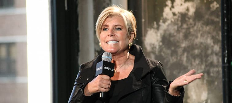 Suze Orman gestures with her hand, talking into a microphone on a stage.