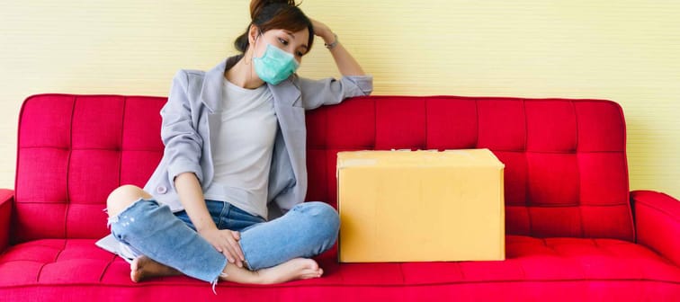Young woman wearing mask sitting on red couch at home with feeling sad and disheartened from company layoff during covid-19, reduction cost. Pandemic disease and impacts every business.