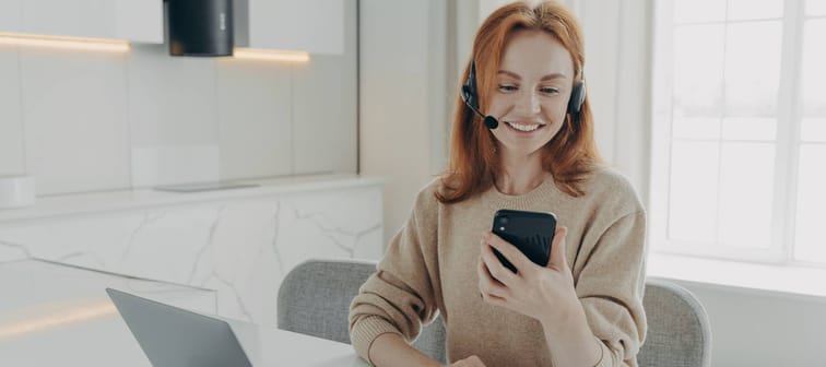Smiling female office worker in headset using smartphone while sitting at her workplace with laptop