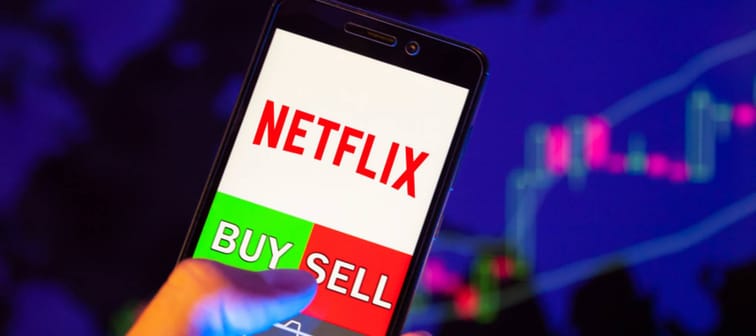 Company logo Netflix on smartphone screen, hand of trader holding mobile phone showing BUY or SELL on background of stock chart