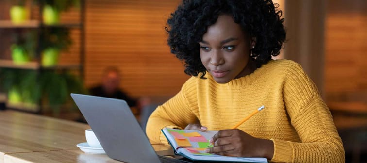 Concentrated black girl browsing job opportunities on laptop at cafe, writing in notepad